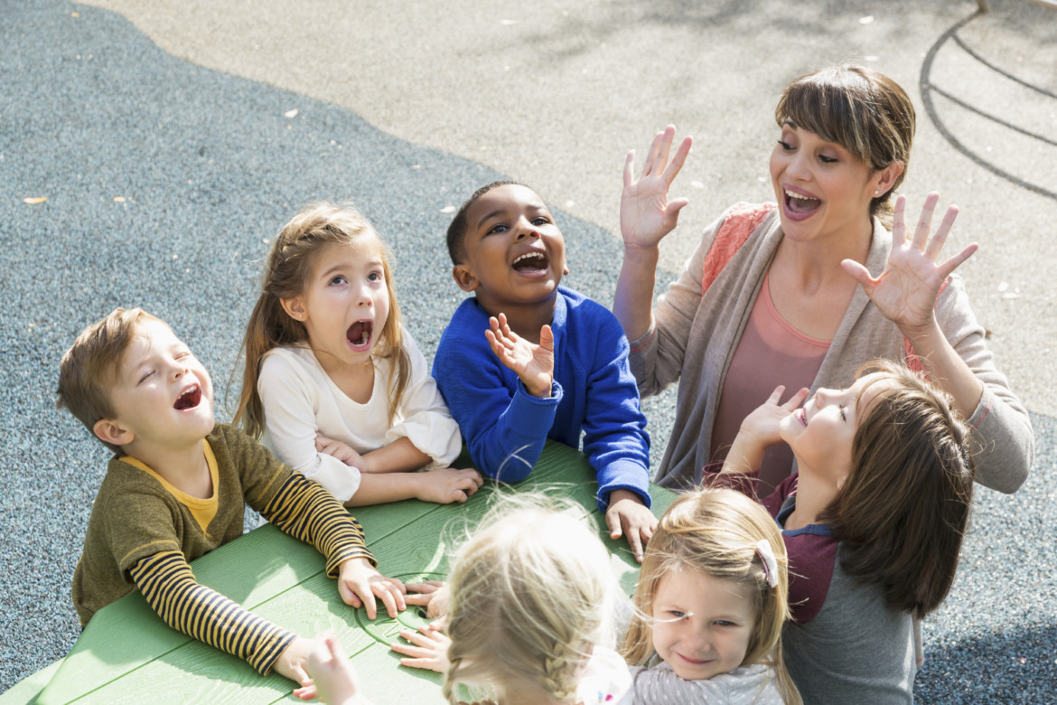 High angle view of a preschool teacher with a multi-ethnic group of children, 4 to 5 years, laughing and having fun. They are sitting at a green table outdoors, mouths open, laughing and shouting.