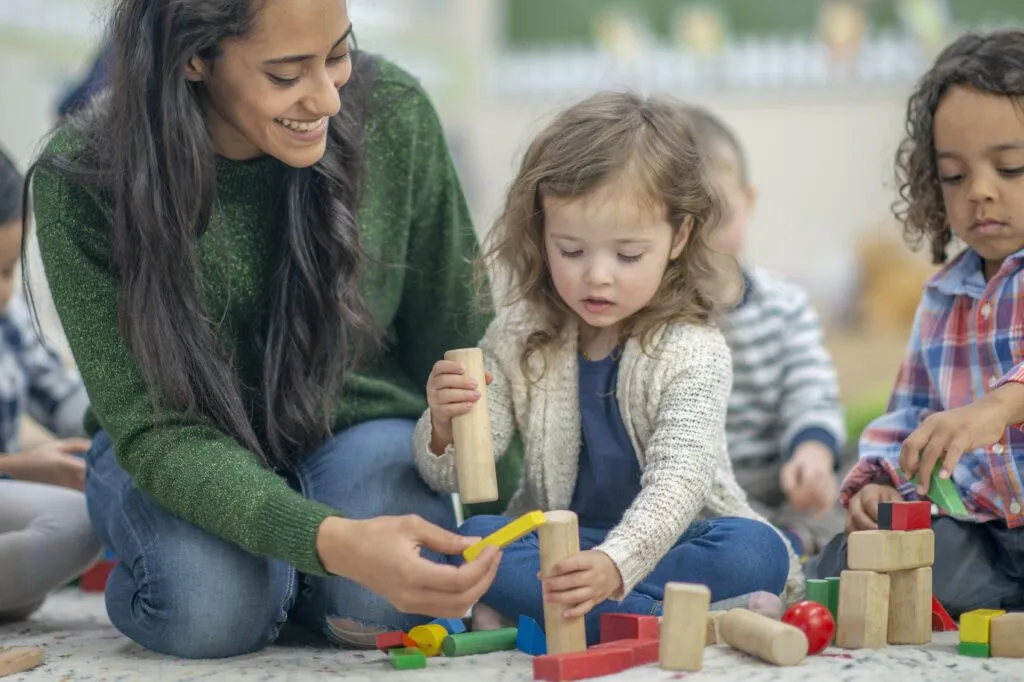 Preschool age children are seated on their classroom floor playing with wooden blocks. Their female teacher is seated next to them and helping one of the children with her blocks.