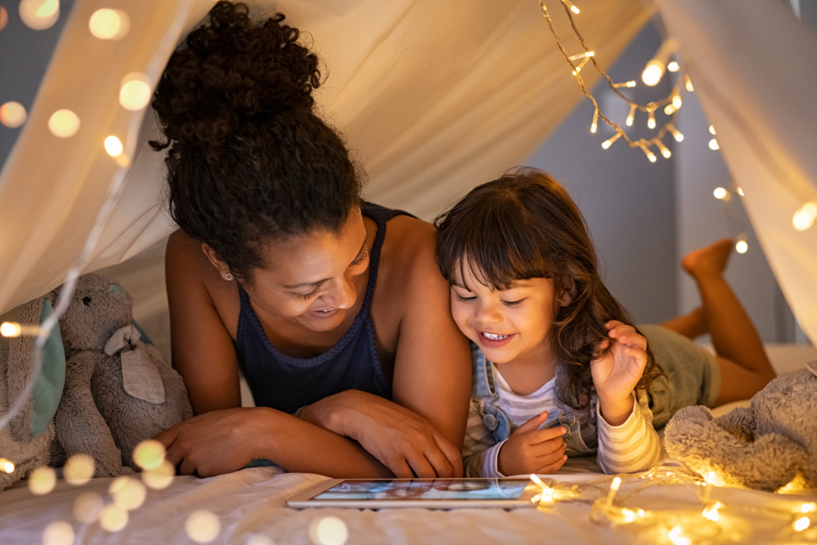African mother and cute smiling girl using digital tablet while lying in illuminted tent in kid bedroom. Cheerful ethnic woman and lovely daughter on video call under a cozy hut. Lovely little girl with mom watching cartoon on digital tablet in bedroom. A mother and preschool age daughter reading together on a tablet.