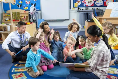 A teacher reading a book aloud as small children sit on the rug listening. Some children have their arms raised. An additional teacher is in the background.