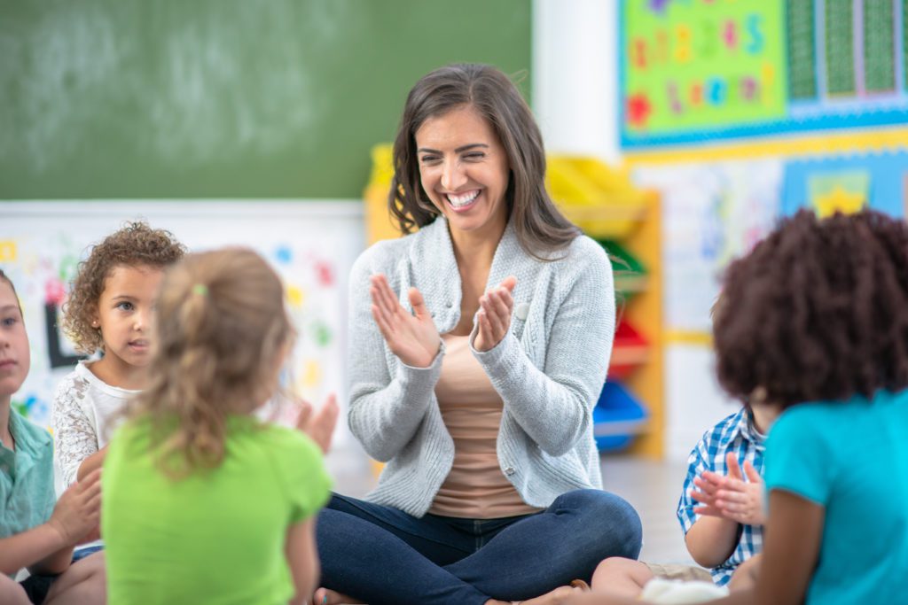 A teacher sits on the floor during small group clapping with children.