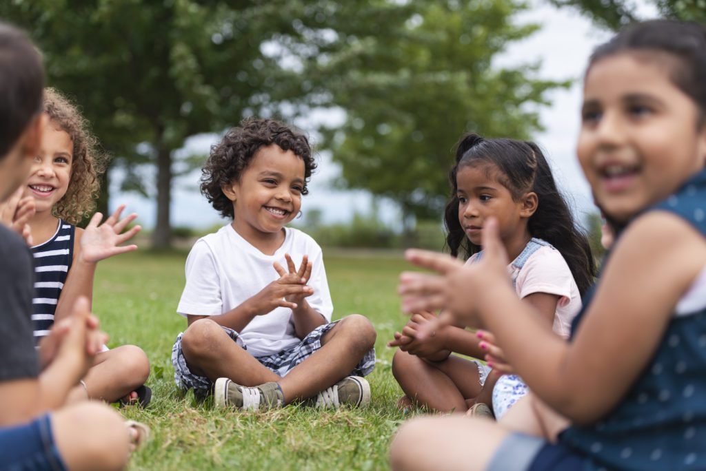 A Group of Young Children are Sitting Together in a Circle, Clapping Outside