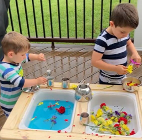 Two boys plays at a water table, one is using the pair of purple scissors.