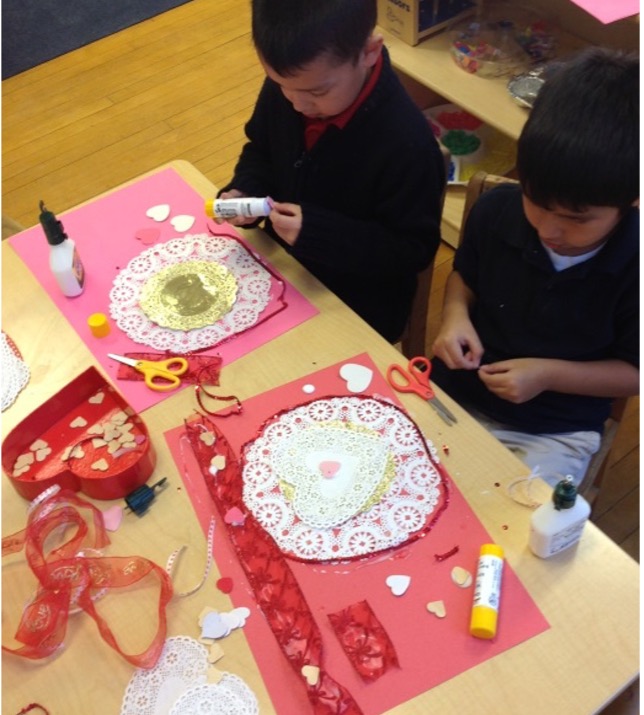 Two young boys create Valentine's Day crafts on large pink and red paper, with a variety of materials including ribbon, doilies, paper hearts, and glue.
