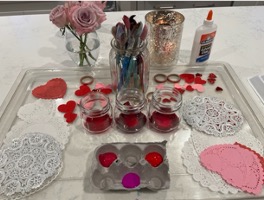 A tray of art materials for Valentine's Day crafts, including glue, flowers, paint brushes, heart stickers, and doily hearts.