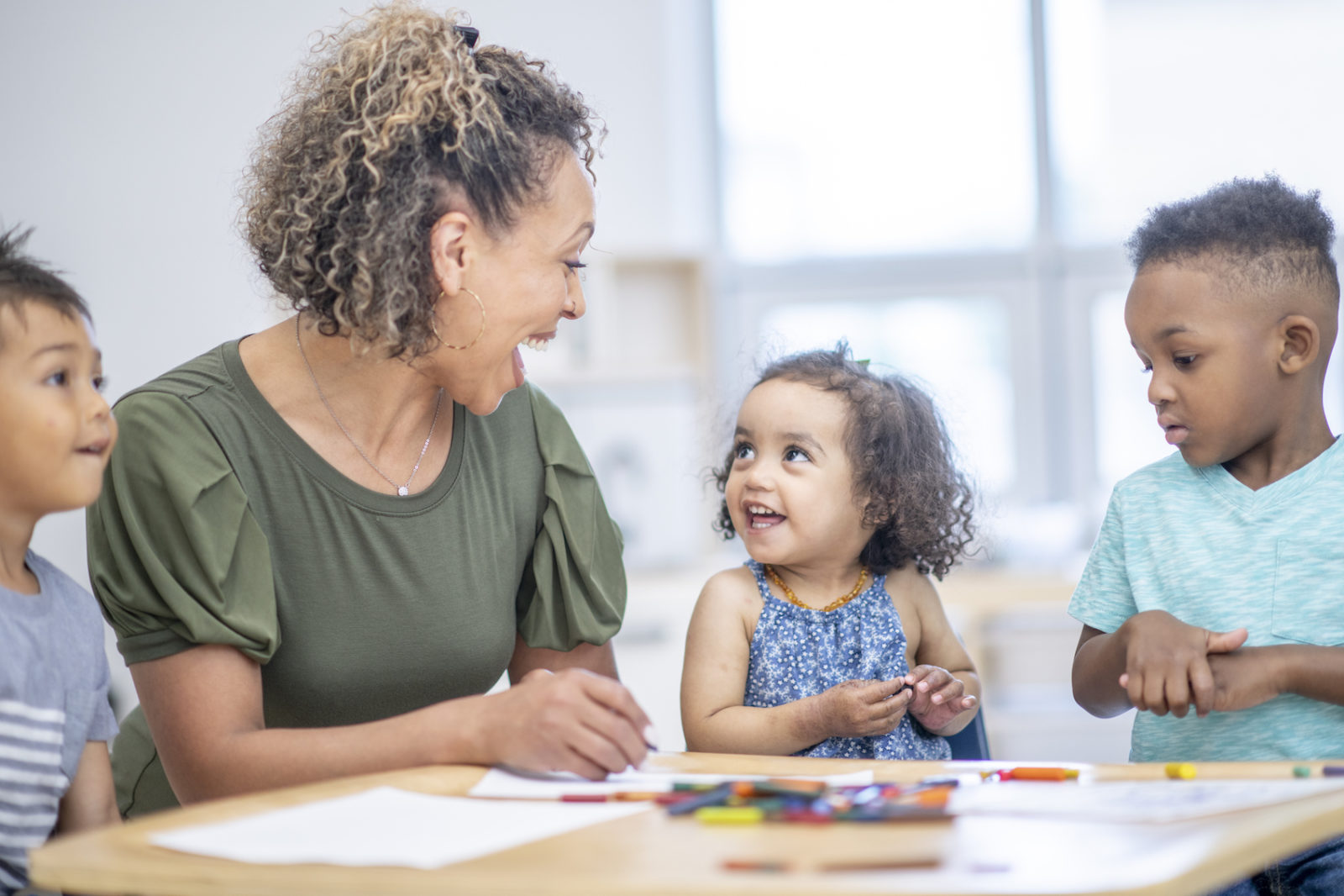 A preschool teacher laughs with her students while sitting at a desk. They are coloring and fully engaged.