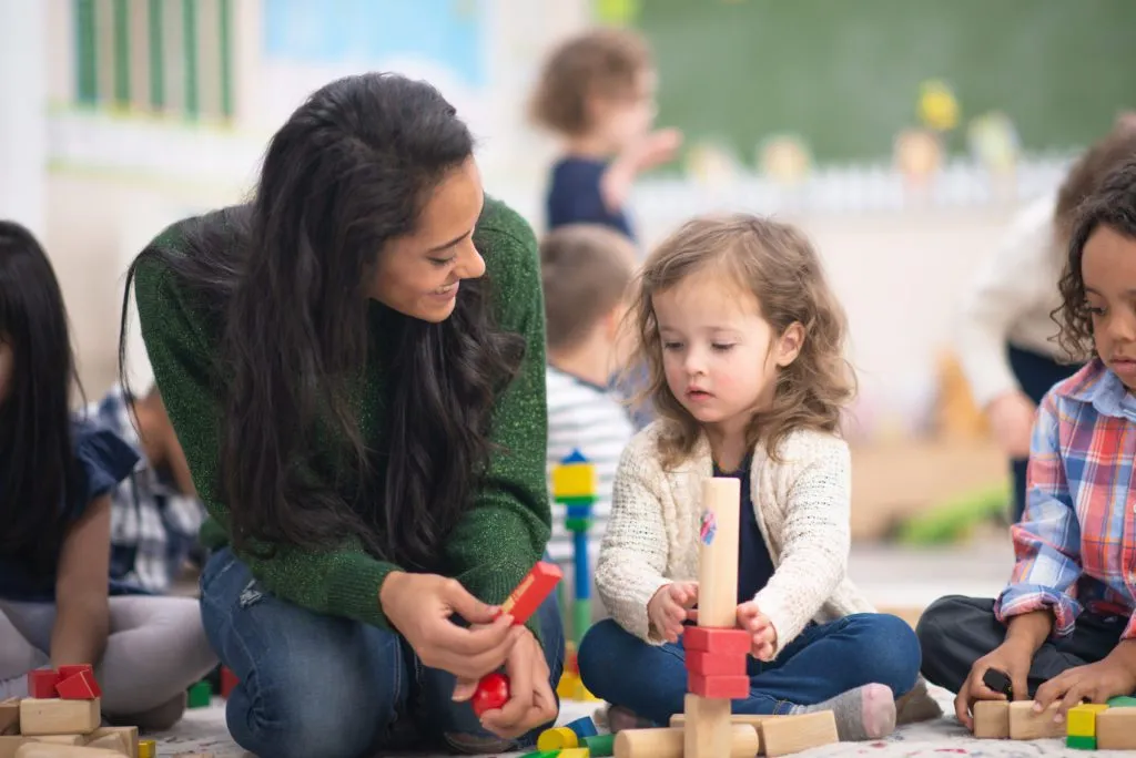 A beautiful teacher holds out a block while she kneels beside her students. Her students are looking at the blocks that they are playing with while sitting on the carpet in a classroom.