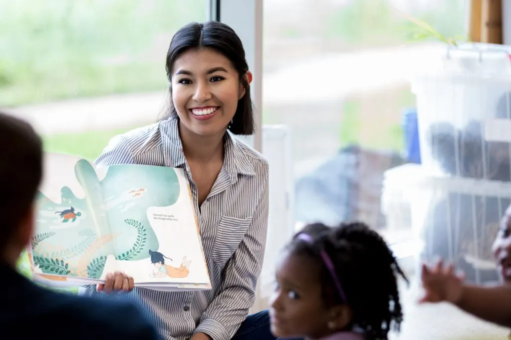 young adult female kindergarten teacher smiles while reading a story book to a group of students.