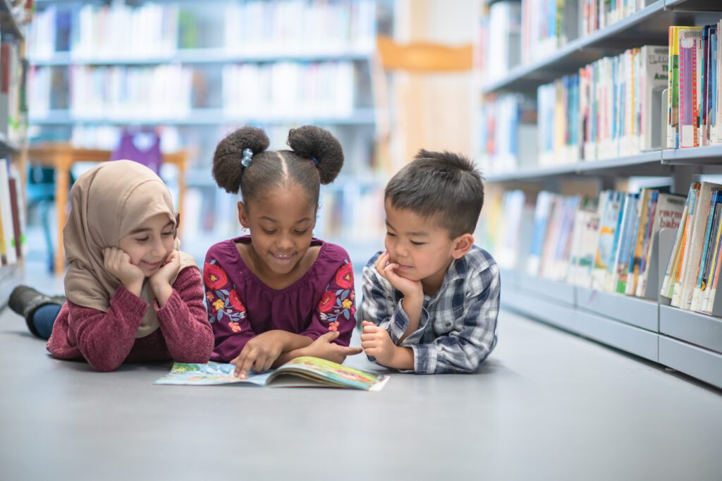 An adorable trio of preschool children and lying on the floor at their school's library as they share a book together.