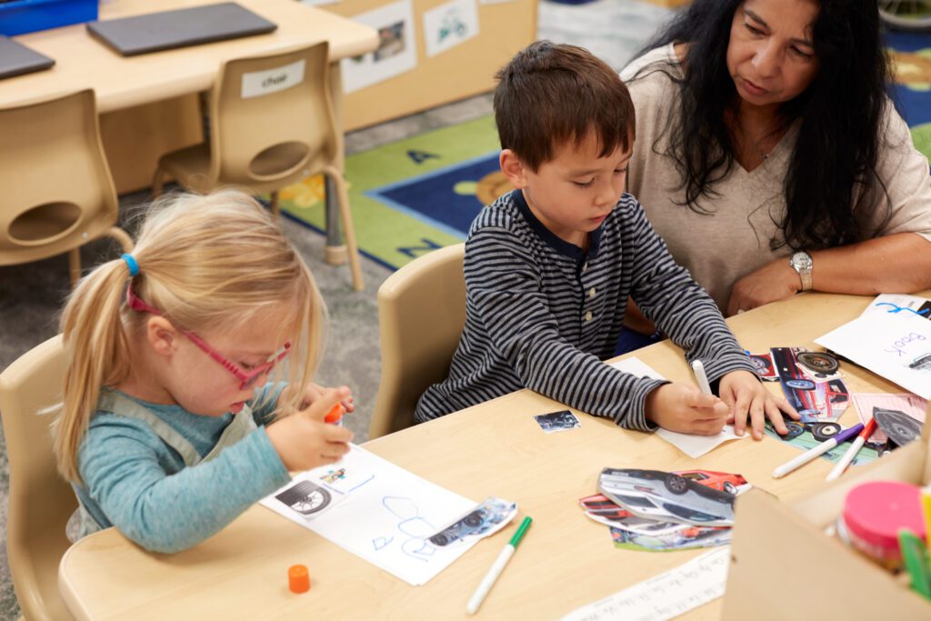 Two pre-k children working on writing and describing what they see at a group table while a teacher looks on nearby.