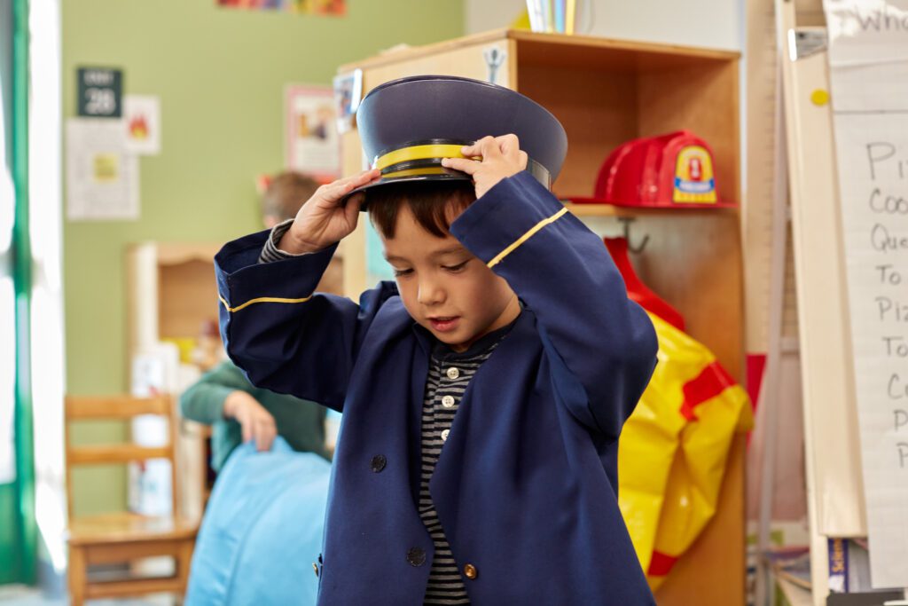 A young boy puts on a conductor coat and hat in the dress up area at school.