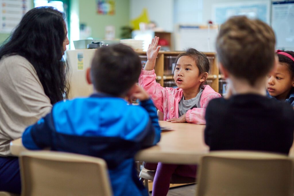 A child raising her hand at a table with her peers and teacher.