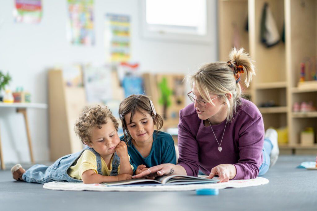 Two children laying on the floor reading a book with the teacher