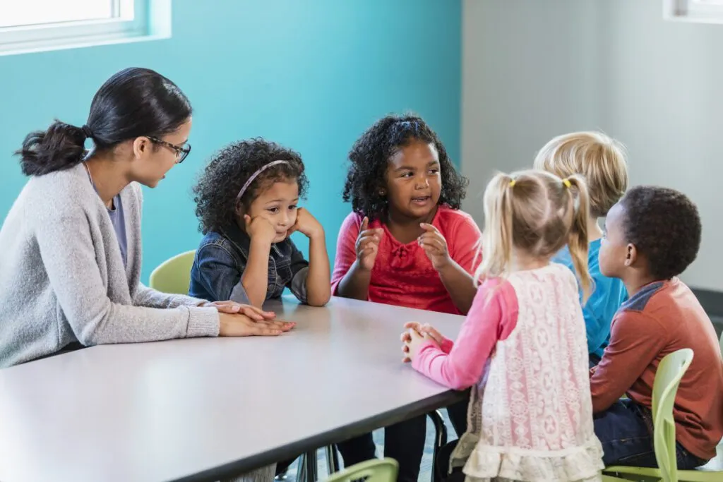 Five students and their teacher sitting at a table talking to eachother